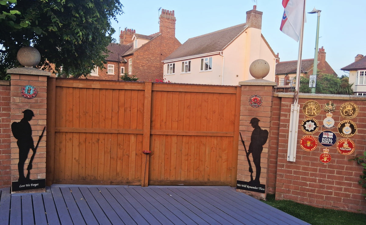Two black wooden soldier memorials beside garden gates, personalised with ‘Lest We Forget’ and ‘We Will Remember Them’.