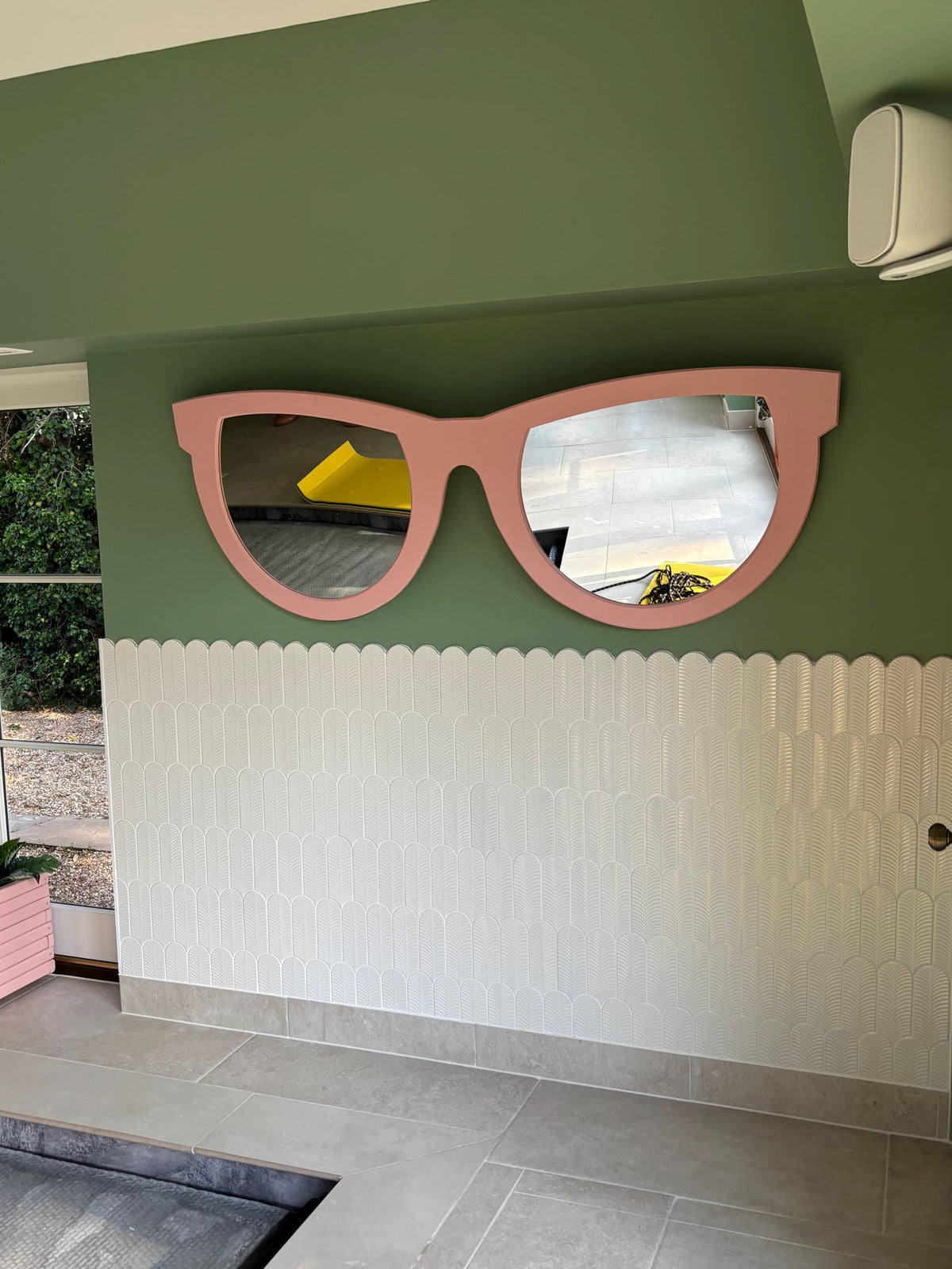 Extra large pink sunglasses-shaped wall mirror mounted on a green wall above white tiles.