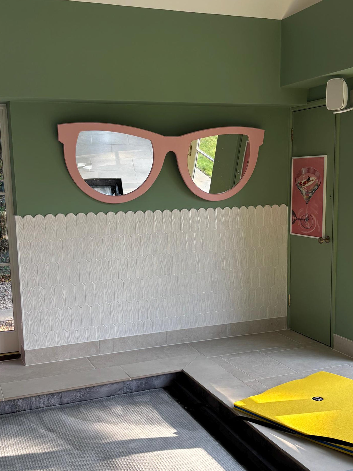 Wide room view of an extra large pink sunglasses-shaped wall mirror above white tiles