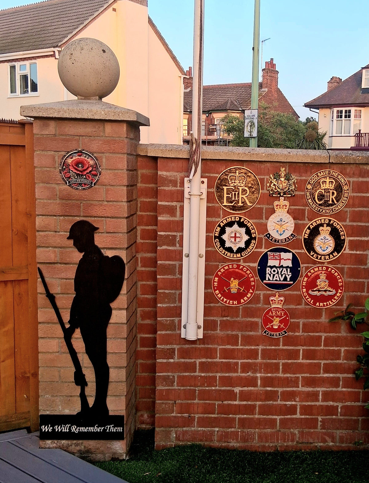 Black wooden soldier memorial with ‘We Will Remember Them’ wording, displayed beside military badges on a brick wall.