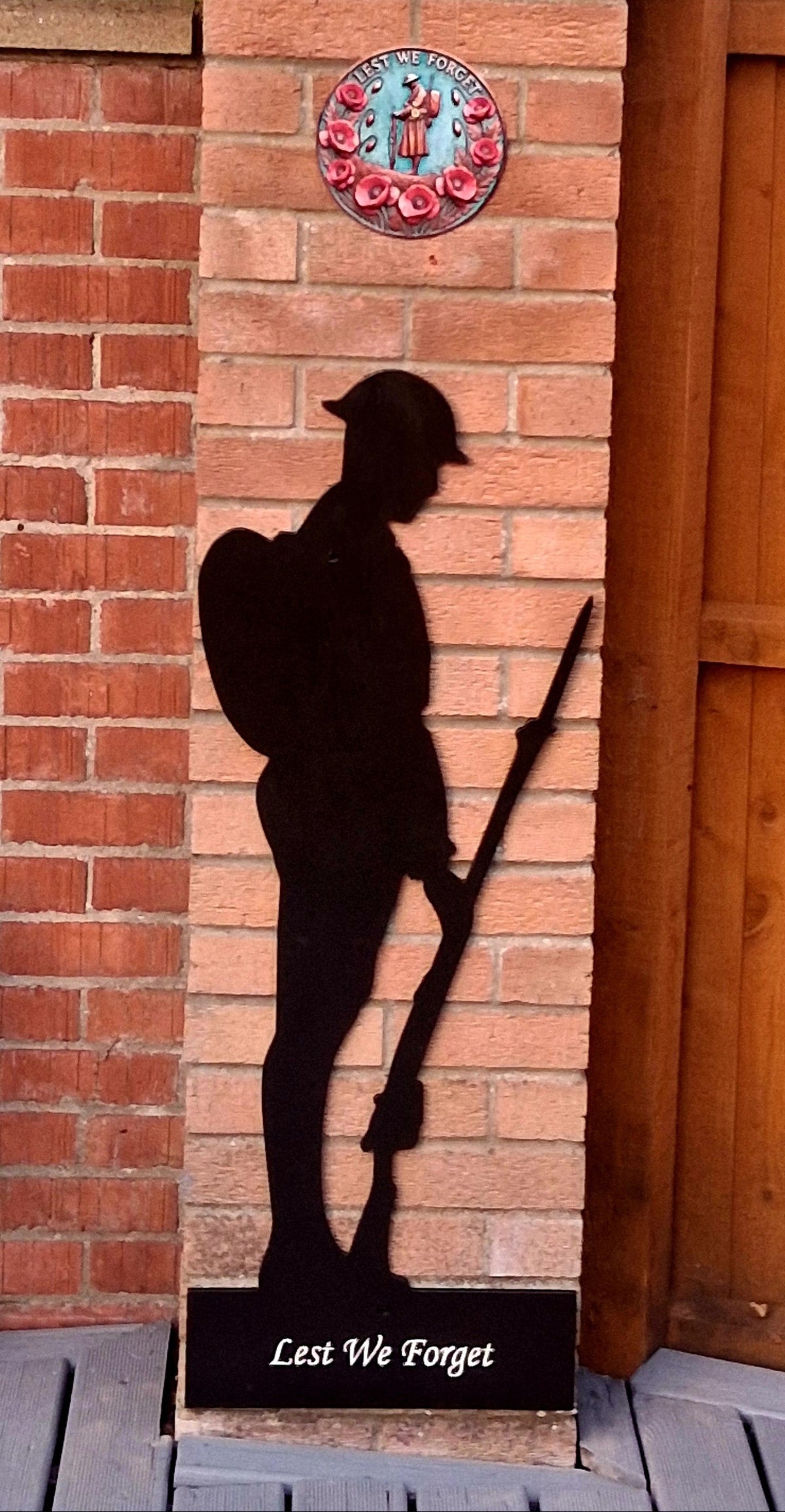 Black wooden soldier memorial against a brick gatepost with ‘Lest We Forget’ wording at the base
