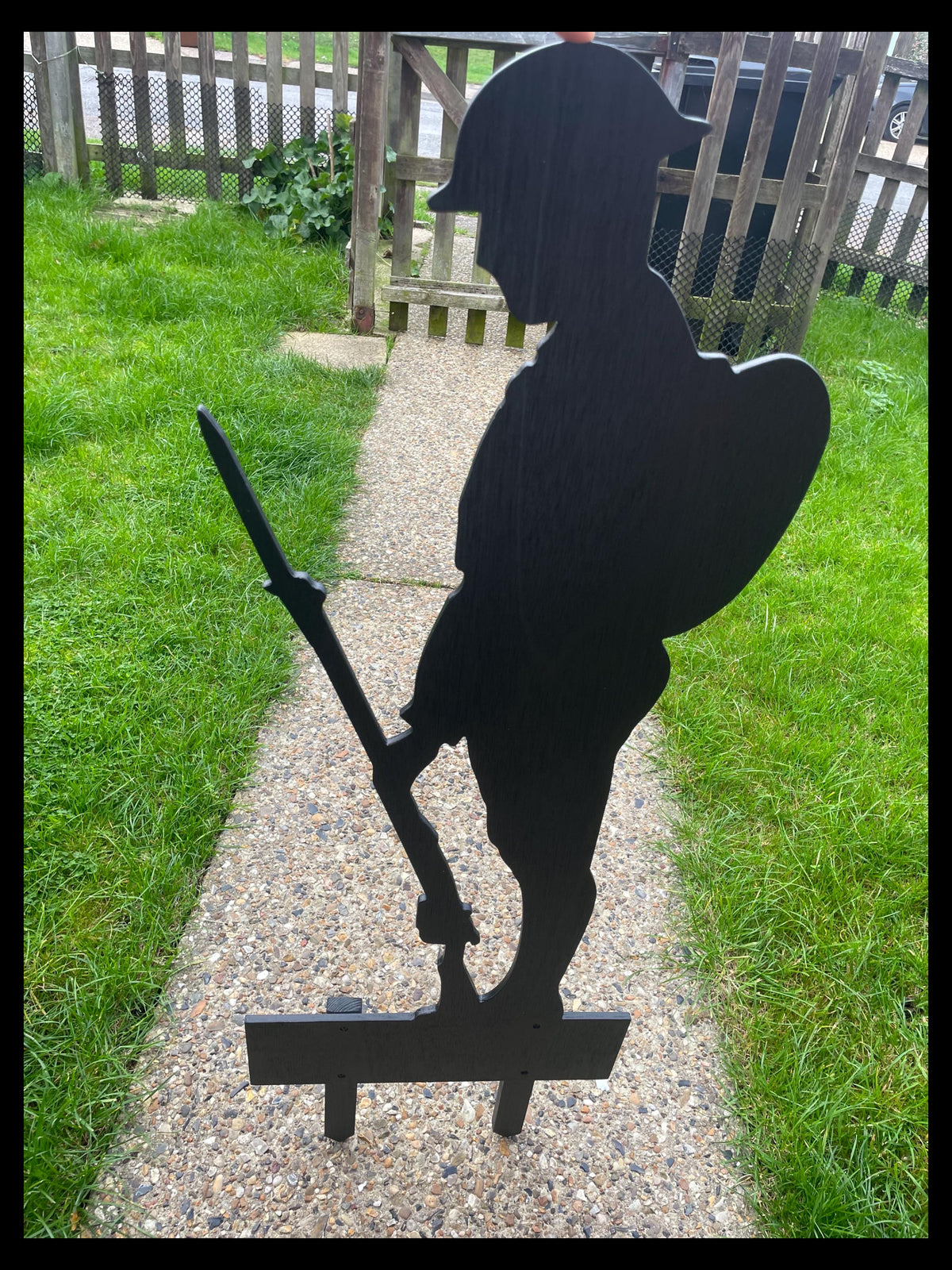 Black wooden soldier memorial silhouette holding a rifle, shown outdoors with two ground stakes.