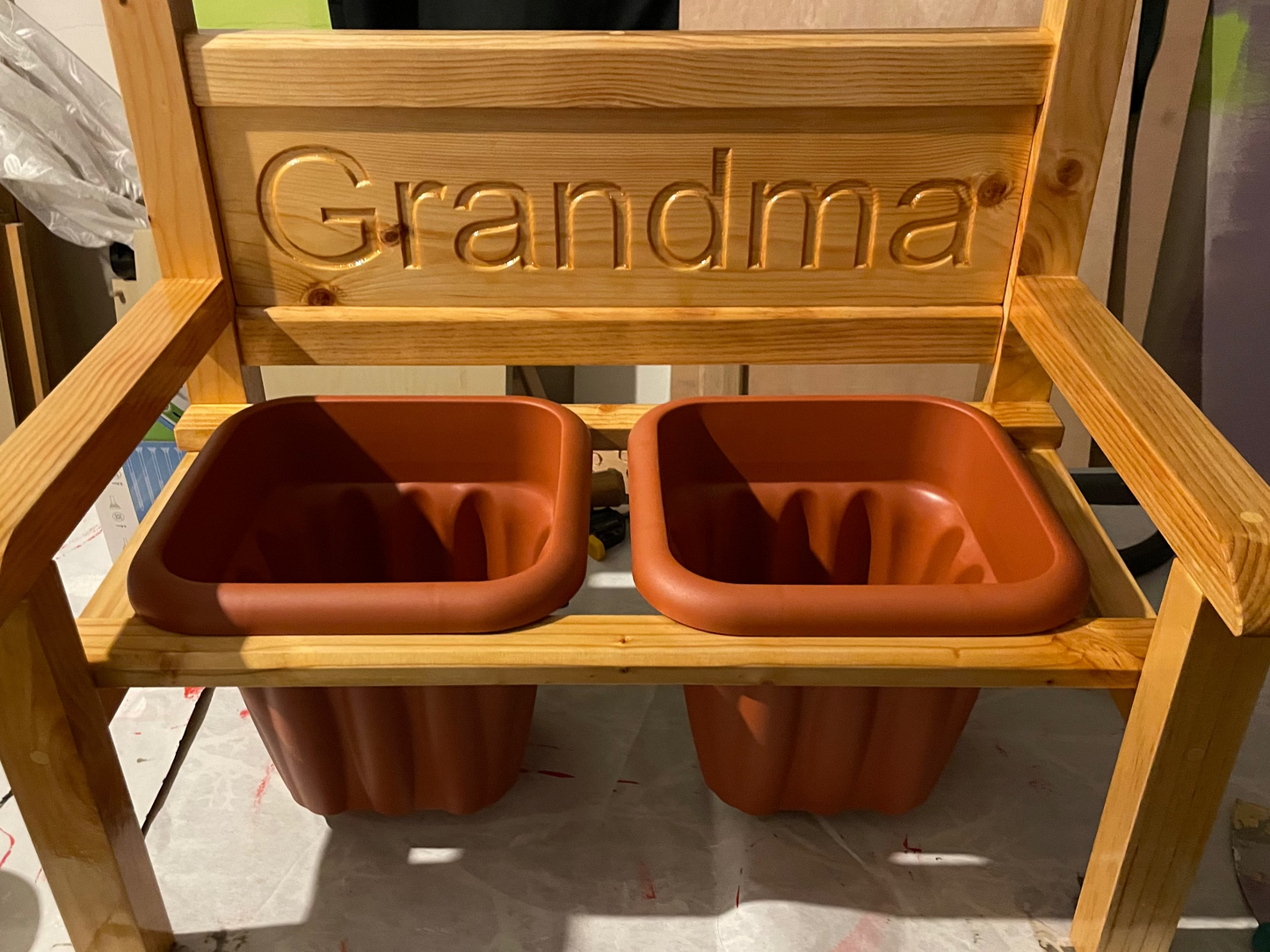 A wooden garden flower bench with the word 'Grandma' engraved, featuring two terracotta planters.