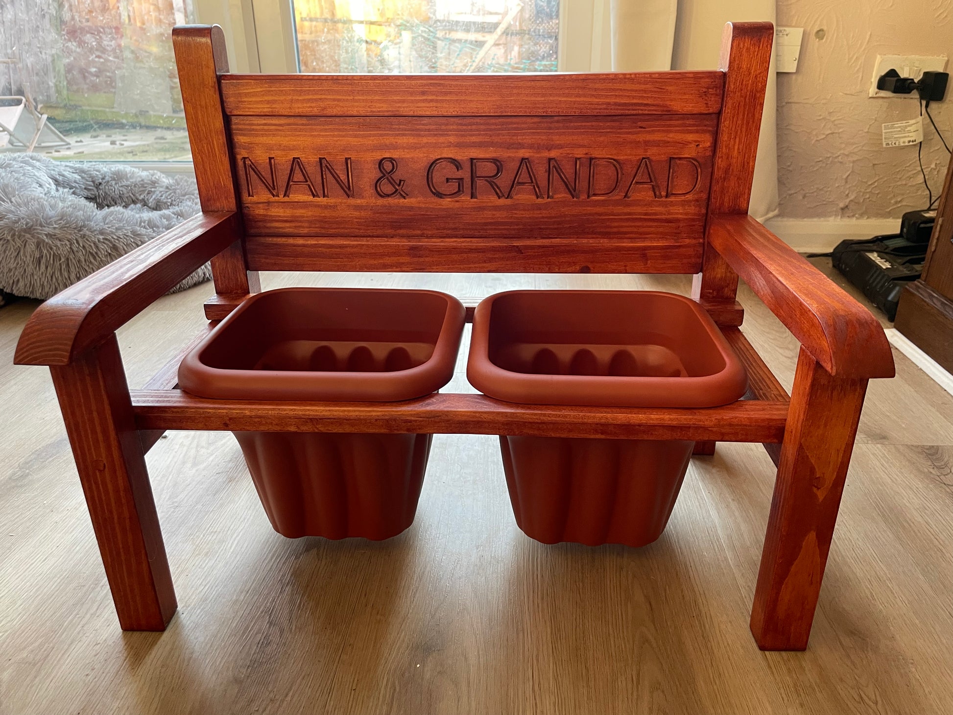 A wooden red stained flower bench with two terracotta pots, The back of the bench is engraved with the words 'NAN & GRANDAD'.