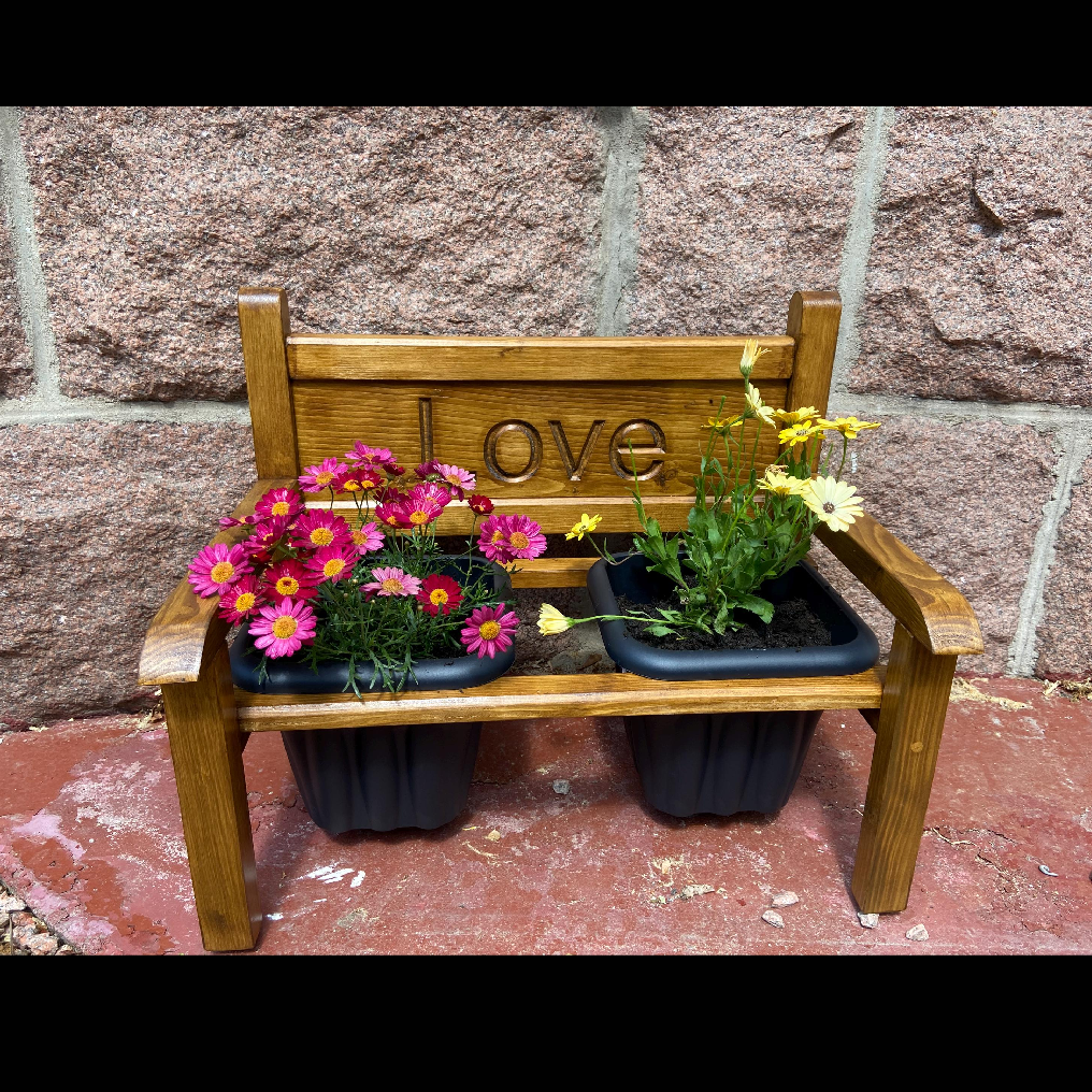 Flower bench with the word 'LOVE' engraved on the back, holding two potted flowers and plants, set against a stone wall.