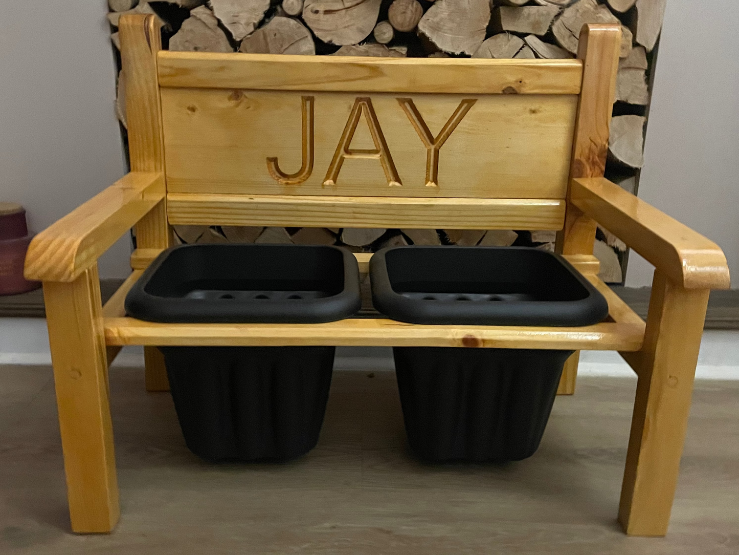 A wooden garden flower bench with the name 'JAY' carved into the backrest, featuring two black plastic planters.