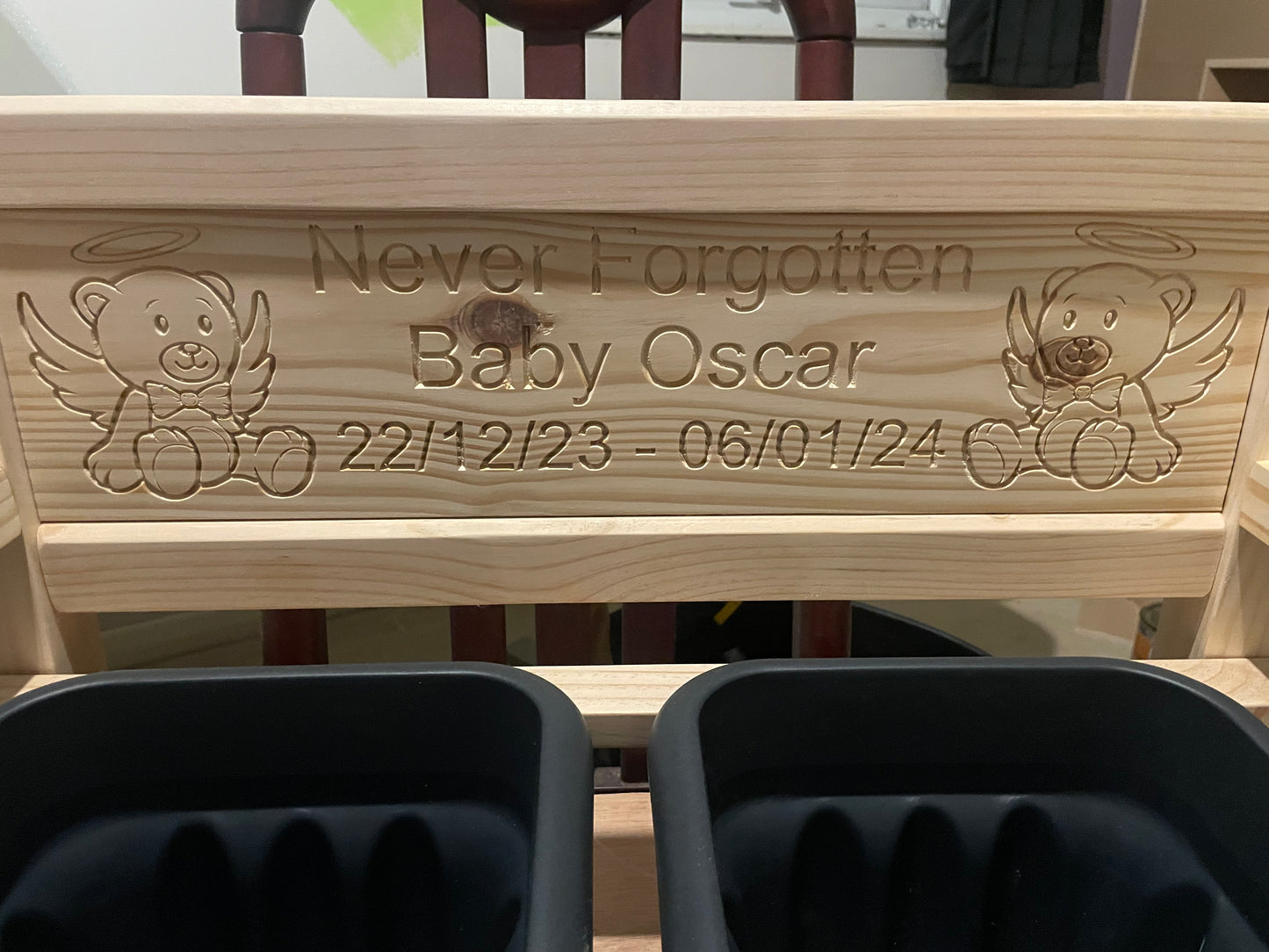 A close-up of a untreated flower bench baby memorial with engraved text reading 'Never Forgotten Baby Oscar' and dates, flanked by angel teddy bear illustrations. Two dark grey flower pots