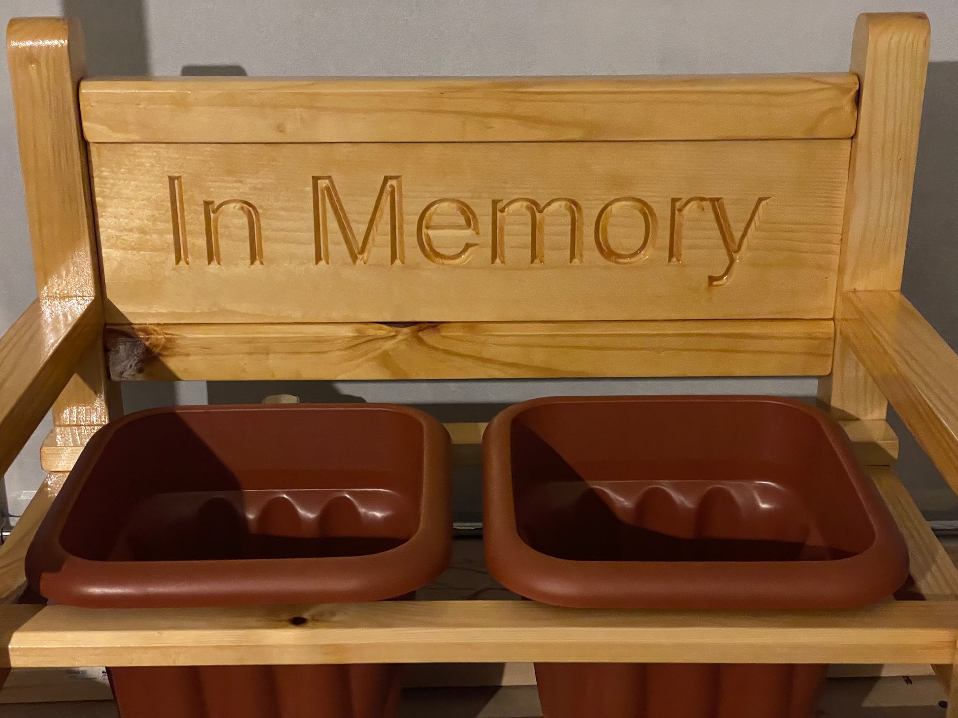 A wooden memorial garden flower bench with 'In Memory' engraved on the backrest, holding two terracotta pots on each side.