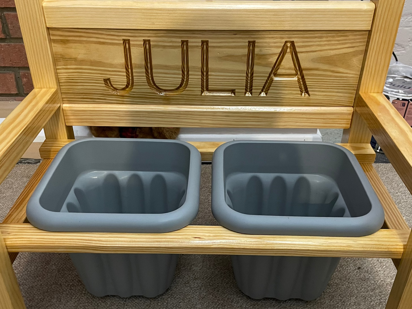 A wooden flower bench with the name 'JULIA' carved into the backrest, featuring two grey pots.