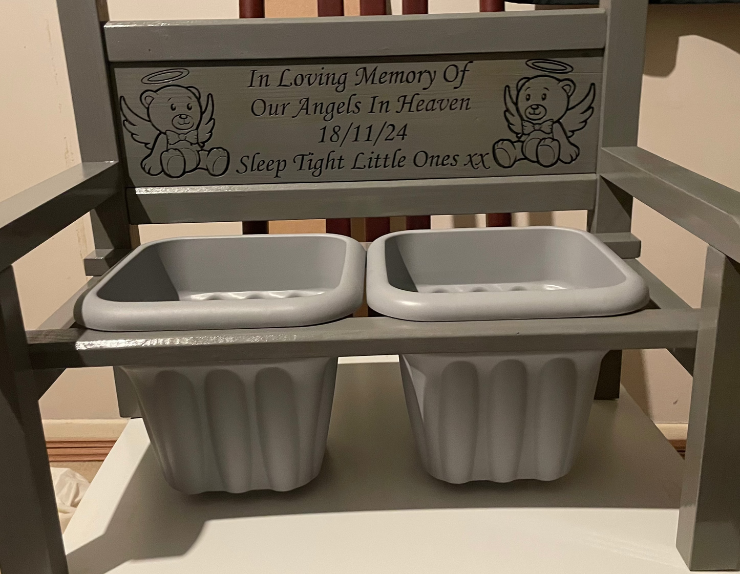 A wooden grey memorial flower bench with two grey flower pot holders, featuring engraved teddy bear illustrations and a remembrance inscription on the backrest.