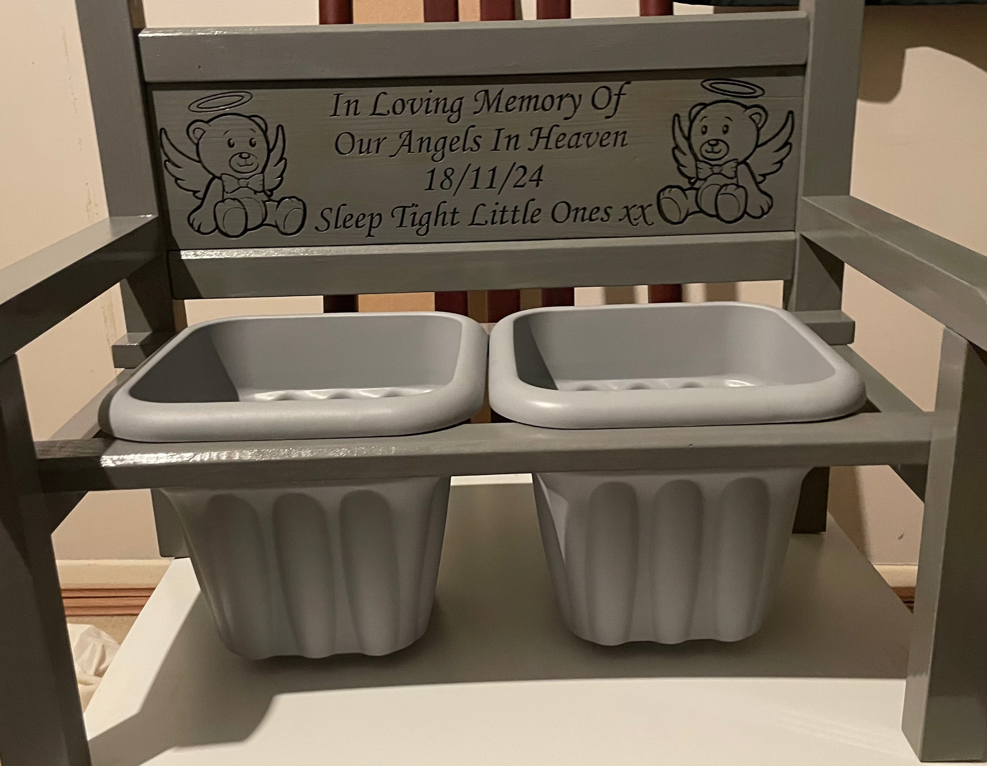 A wooden grey memorial flower bench with two grey flower pot holders, featuring engraved teddy bear illustrations and a remembrance inscription on the backrest.