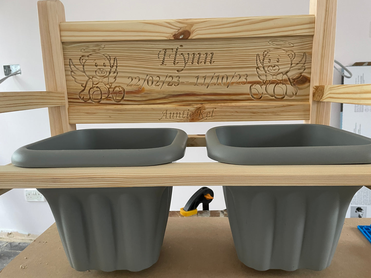 A untreated wooden flower bench with two grey pots, featuring an engraved wooden backrest with the name 'Flynn' and angel illustrations. with engraving on the rails